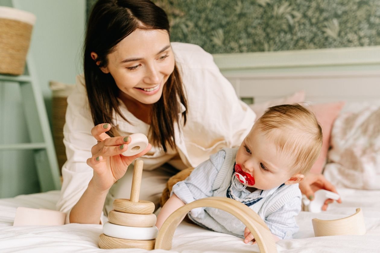 Mother and baby boy playing together with a wooden toy on a cosy bed